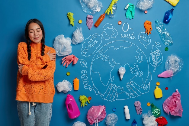 Foto gratuita complacida joven activista se abraza a sí misma, se siente cómoda, posa contra la pared azul con desechos plásticos alrededor del mundo, lucha contra la contaminación ambiental