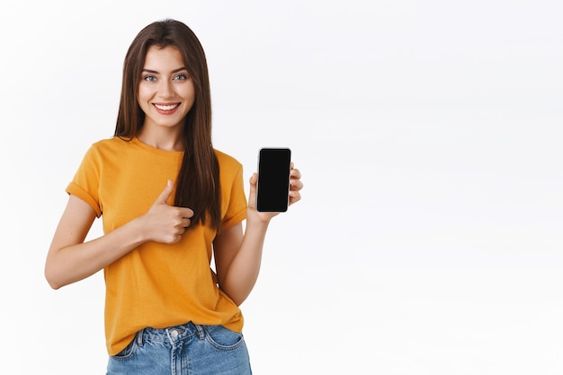 Foto gratuita complacida, satisfecha, atractiva y elegante chica con camiseta amarilla que muestra la aplicación de teléfono inteligente con pantalla de teléfono móvil, hace el pulgar hacia arriba y sonríe en acuerdo de aprobación, da recomendación