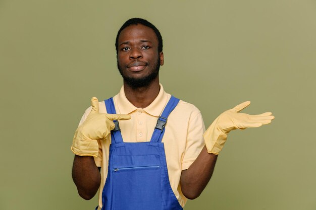 Complacido fingiendo sostener y apuntando a algo joven limpiador afroamericano en uniforme con guantes aislados en fondo verde
