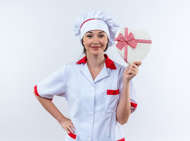 Complacido joven cocinera vistiendo uniforme de chef sosteniendo una caja en forma de corazón poniendo la mano en la cadera aislado sobre fondo blanco.