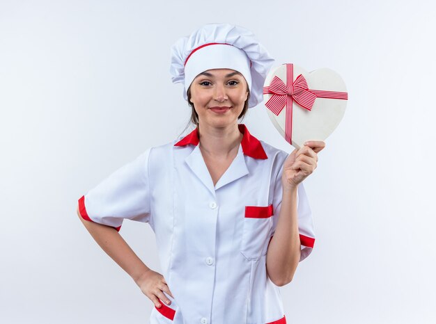 Complacido joven cocinera vistiendo uniforme de chef sosteniendo una caja en forma de corazón poniendo la mano en la cadera aislado sobre fondo blanco.