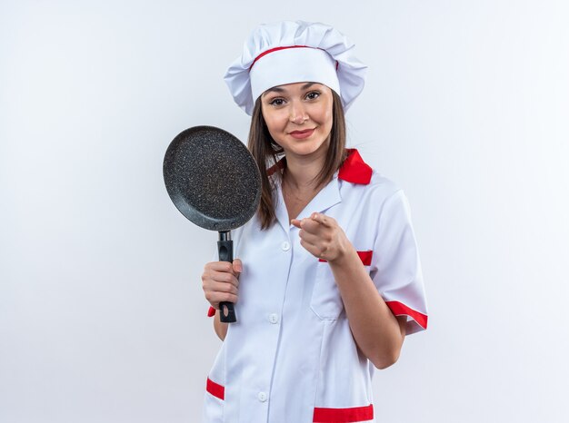 Complacido joven cocinera vistiendo uniforme de chef sosteniendo puntos de sartén en la cámara aislada sobre fondo blanco.