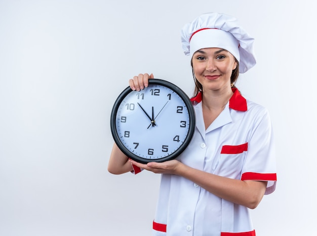 Complacido joven cocinera vistiendo uniforme de chef sosteniendo el reloj de pared aislado sobre fondo blanco.