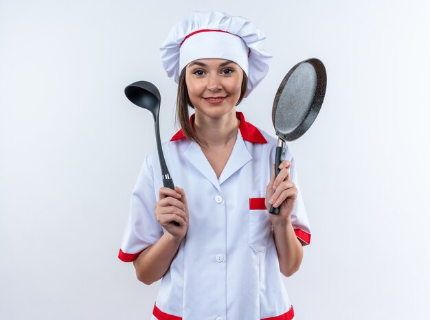 Complacido joven cocinera vistiendo uniforme de chef sosteniendo una sartén con cucharón aislado sobre fondo blanco.