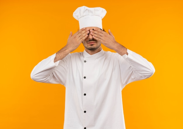 Complacido joven cocinero con uniforme de chef y gafas cubriendo los ojos con las manos aisladas en la pared amarilla