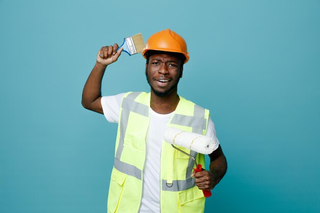 Complacido joven constructor afroamericano en uniforme sosteniendo cepillo de rodillos con pincel aislado sobre fondo azul.