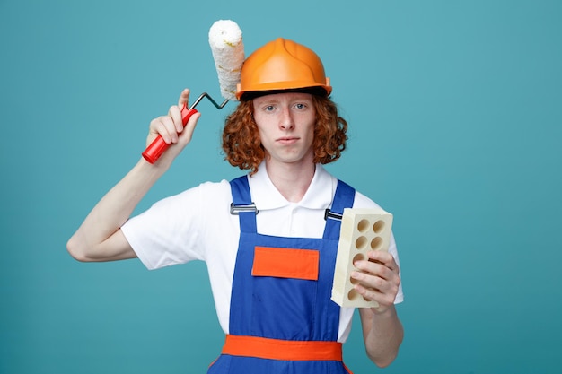 Complacido joven constructor hombre en uniforme sosteniendo cepillo de rodillos y ladrillo aislado sobre fondo azul.