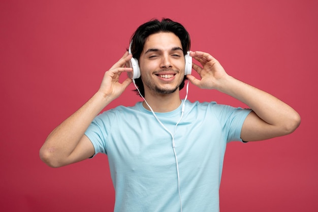 complacido joven guapo usando y agarrando auriculares mirando a la cámara sonriendo aislado sobre fondo rojo.