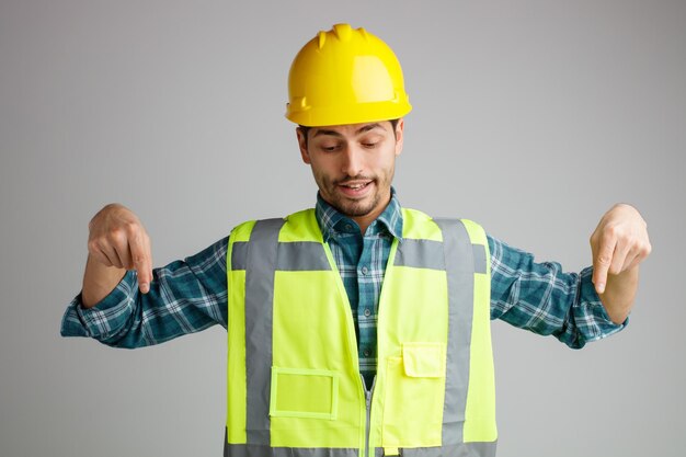 Complacido joven ingeniero masculino con casco de seguridad y uniforme mirando y apuntando hacia abajo aislado sobre fondo blanco.