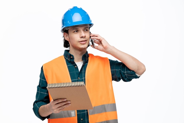 Complacido joven ingeniero masculino usando casco de seguridad y chaleco de seguridad sosteniendo bloc de notas mirando al lado hablando por teléfono aislado sobre fondo blanco.