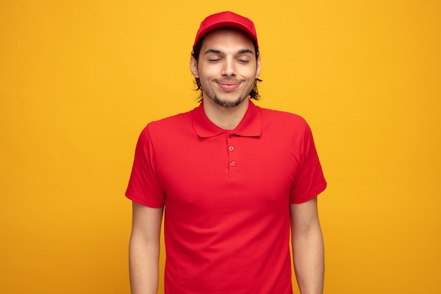 complacido joven repartidor con uniforme y gorra sonriendo con los ojos cerrados aislado sobre fondo amarillo