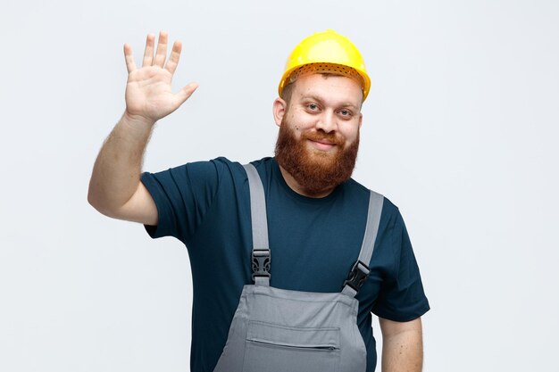 Complacido joven trabajador de la construcción con casco de seguridad y uniforme mirando a la cámara mostrando hola gesto aislado sobre fondo blanco.