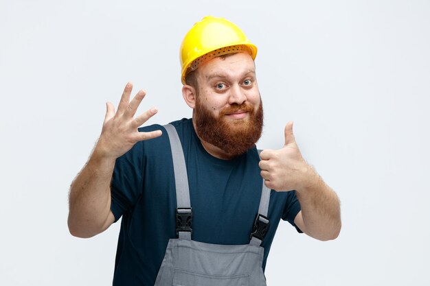 Complacido joven trabajador de la construcción con casco de seguridad y uniforme mirando a la cámara que muestra cinco con la mano y el pulgar hacia arriba aislado sobre fondo blanco.