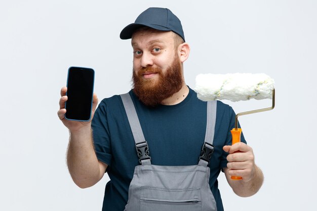 Complacido joven trabajador de la construcción con gorra y uniforme sosteniendo un rodillo de pintura mirando a la cámara mostrando el teléfono móvil a la cámara aislada en el fondo blanco