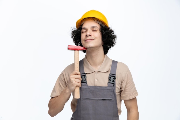 Complacido joven trabajador de la construcción vistiendo uniforme y casco de seguridad tocando la barbilla con un martillo con los ojos cerrados aislado sobre fondo blanco.