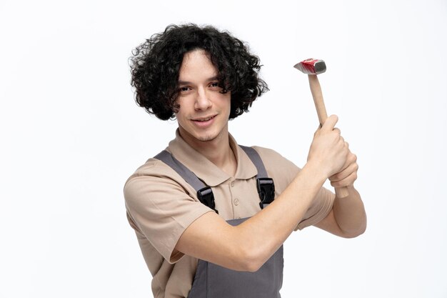 Complacido joven trabajador de la construcción vistiendo uniforme mirando a la cámara sosteniendo un martillo con ambas manos aislado sobre fondo blanco.