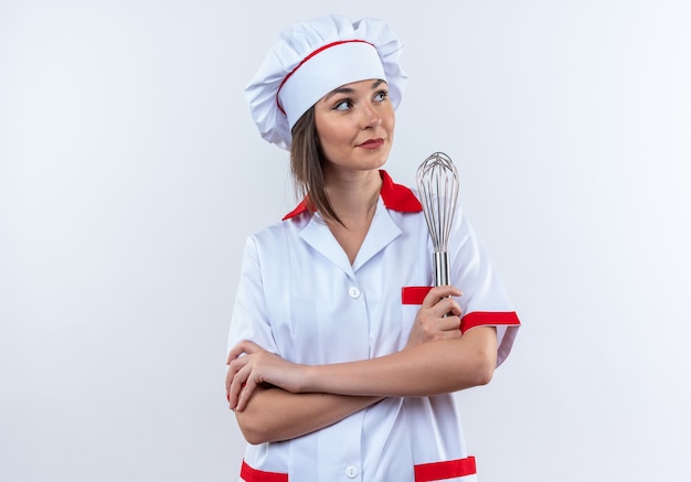 Complacido mirando lado joven cocinera vistiendo uniforme de chef sosteniendo batidor cruzando las manos aisladas sobre fondo blanco