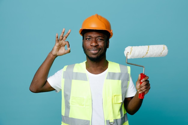 Complacido mostrando buen gesto joven constructor afroamericano en uniforme sosteniendo cepillo de rodillos aislado sobre fondo azul.