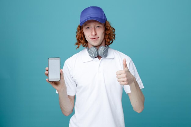 Complacido mostrando los pulgares hacia arriba joven apuesto con gorra usando auriculares sosteniendo el teléfono aislado en el fondo azul