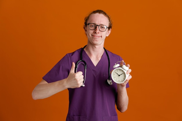 complacido mostrando Thumbs up sosteniendo despertador joven médico masculino vistiendo uniforme con estetoscopio aislado sobre fondo naranja
