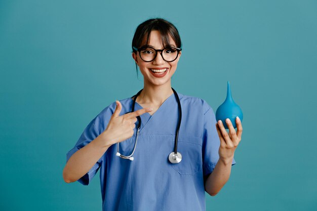 Complacido mostrando Thumbs up sosteniendo enemas joven doctora vistiendo uniforme fith estetoscopio aislado sobre fondo azul.