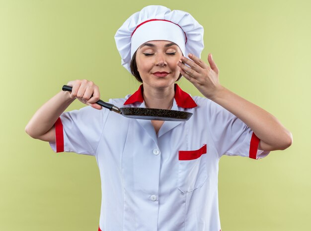Complacido con los ojos cerrados joven cocinera vistiendo uniforme de chef sosteniendo y oliendo sartén aislado en la pared verde oliva