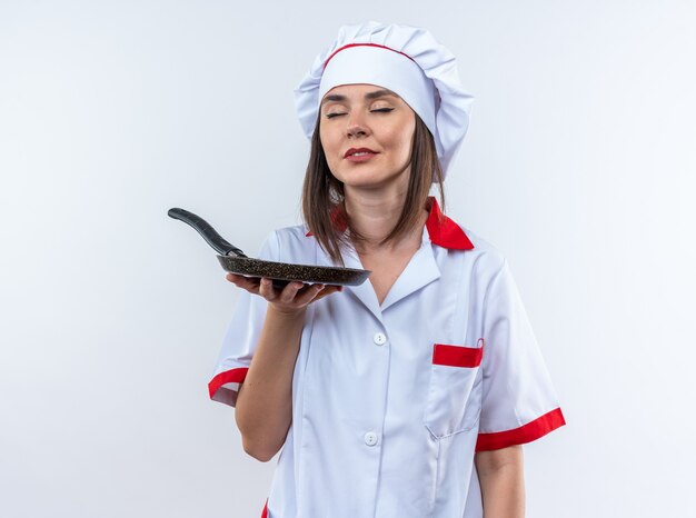 Complacido con los ojos cerrados joven cocinera vistiendo uniforme de chef sosteniendo una sartén aislado en la pared blanca