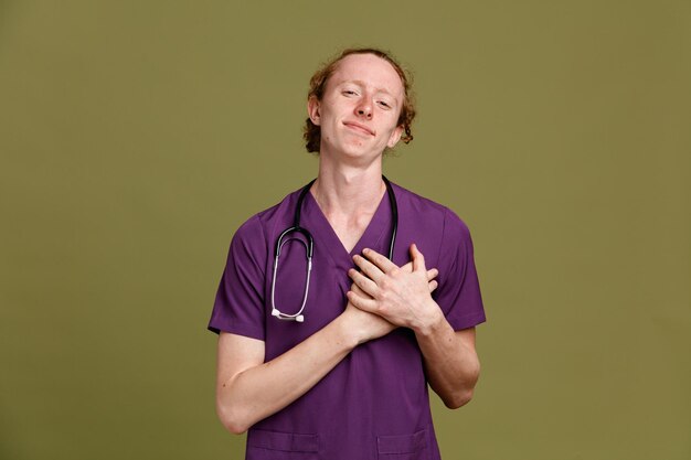 Complacido poniendo la mano en el corazón joven médico masculino vistiendo uniforme con estetoscopio aislado sobre fondo verde