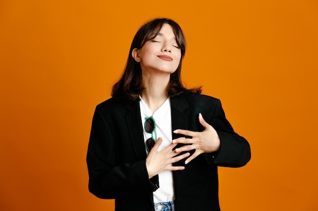 Complacido poniendo las manos en el corazón joven hermosa mujer vistiendo chaqueta negra aislado sobre fondo naranja