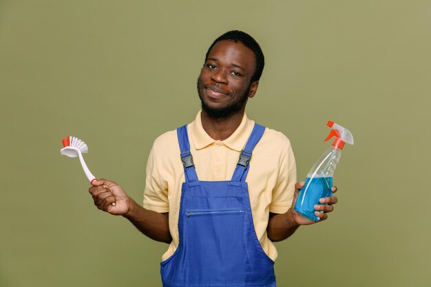 Complacido sosteniendo agente de limpieza con cepillo de limpieza joven limpiador afroamericano en uniforme con guantes aislado sobre fondo verde