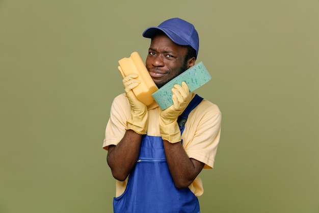 Complacido sosteniendo esponjas de limpieza alrededor de la cara joven limpiador afroamericano en uniforme con guantes aislados en fondo verde