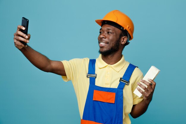 Complacido sosteniendo ladrillo joven constructor afroamericano en uniforme tome un selfie aislado sobre fondo azul.