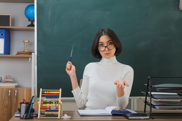 complacido sosteniendo la mano a la cámara joven maestra con gafas puntos con puntero en la pizarra sentada en el escritorio con herramientas escolares en el aula