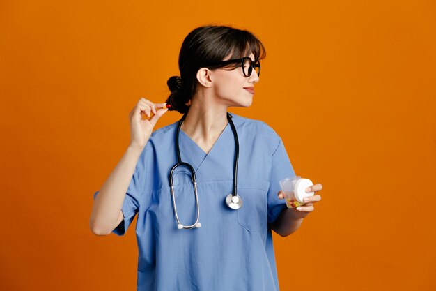 complacido sosteniendo la píldora y el recipiente de la píldora joven doctora vistiendo uniforme fith estetoscopio aislado sobre fondo naranja