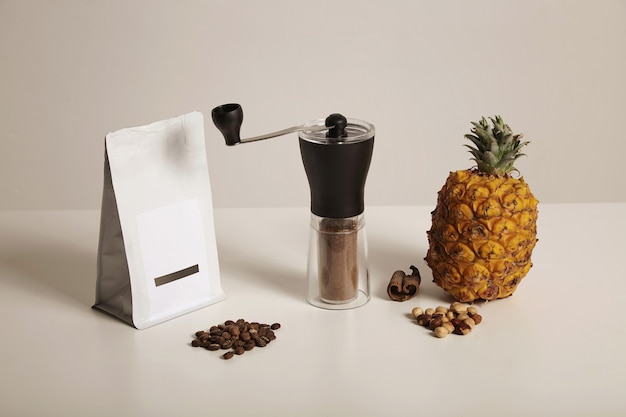 Una composición de café recién molido en un molinillo manual, bolsa de granos de café, nueces, piña y canela en blanco