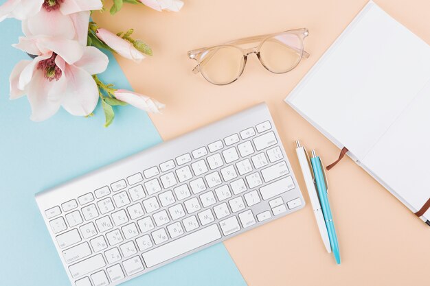 Composición de cuaderno, teclado, lentes, flores y bolígrafos.