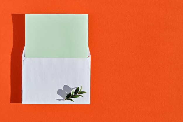 Composición mínima con sobre blanco, tarjeta verde en blanco y hojas verdes sobre fondo naranja quemado, vista superior con espacio de copia. Mockup con sobre y tarjeta en blanco, plano