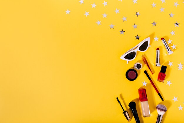 Composición de pinceles de maquillaje, base tonal, delineador de ojos, lápiz labial, rímel y elegantes gafas de sol en una pared naranja con estrellas plateadas