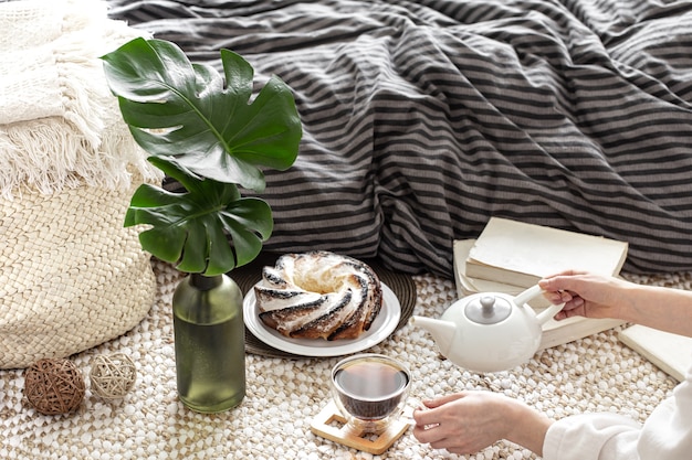 Composición de una taza de té, cupcakes caseros y hojas decorativas en un jarrón con el telón de fondo de una cama acogedora.