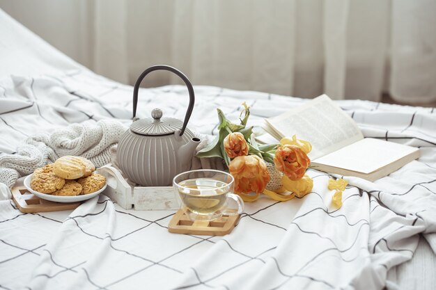 Composición con una taza de té, una tetera, un ramo de tulipanes y galletas en la cama