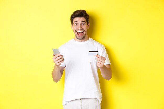 Comprador masculino feliz que sostiene el teléfono inteligente y la tarjeta de crédito, concepto de compras en línea en internet, de pie sobre fondo amarillo.