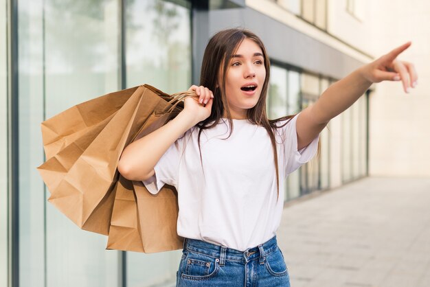 Comprador sorprendido abriendo la boca sosteniendo bolsas de compras viendo ofertas especiales en las tiendas y apuntando en la calle