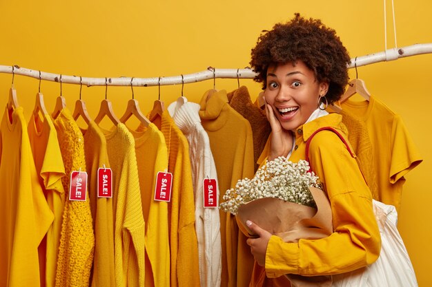 Compradora de mujer de pelo rizado alegre elige ropa a la venta colgada en perchas, lleva bolsa, posa con ramo, aislado sobre fondo amarillo.