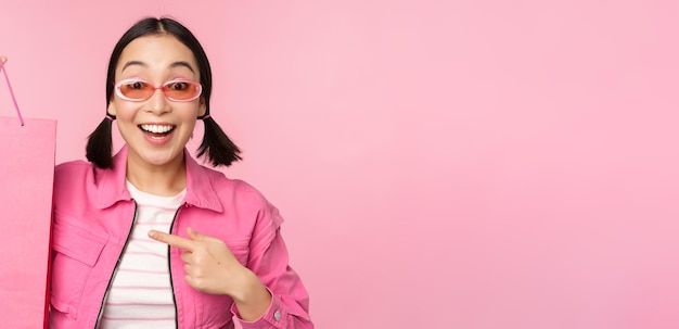 Compras Elegante chica asiática con gafas de sol mostrando una bolsa de la tienda y sonriendo recomendando promoción de venta en la tienda de pie sobre fondo rosa