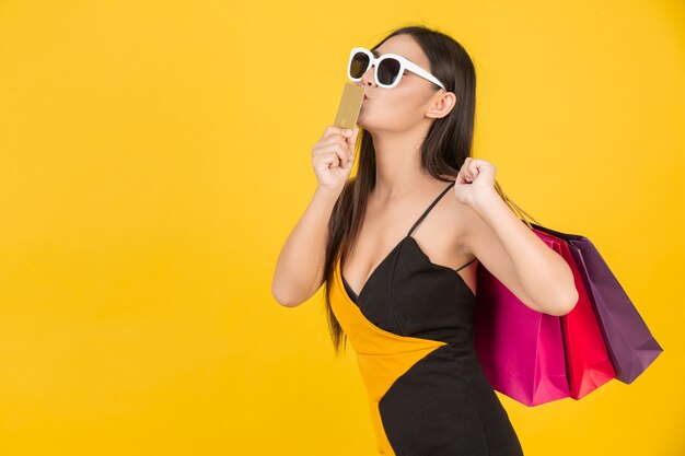 Compras hermosa mujer con gafas con una tarjeta de crédito de oro con una bolsa de papel de colores en un amarillo.