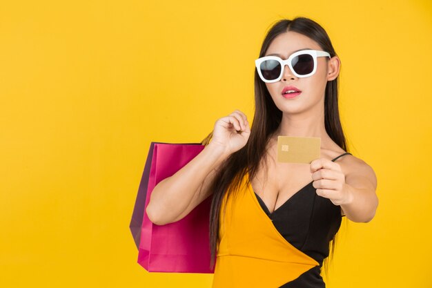 Compras hermosa mujer con gafas con una tarjeta de crédito de oro con una bolsa de papel de colores en un amarillo.