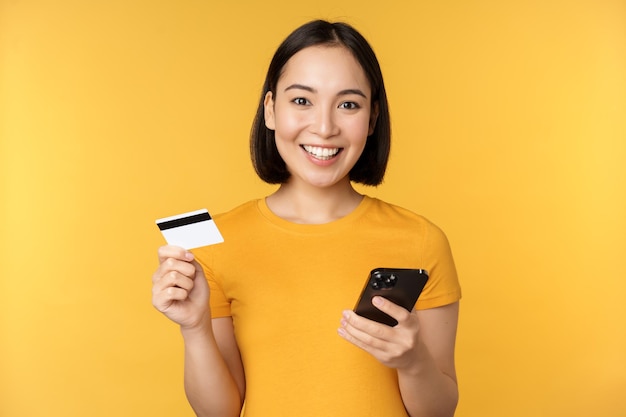 Compras en línea Alegre niña asiática con tarjeta de crédito y orden de pago de teléfono inteligente con teléfono móvil de pie sobre fondo amarillo