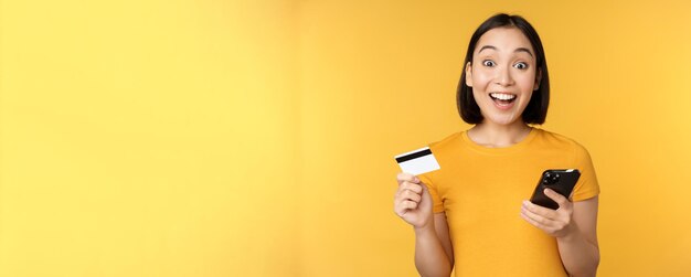 Compras en línea Alegre niña asiática con tarjeta de crédito y orden de pago de teléfono inteligente con teléfono móvil de pie sobre fondo amarillo