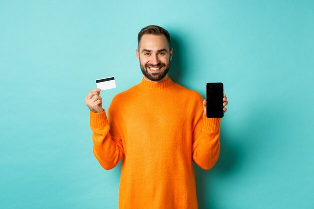 Las compras en línea. Chico atractivo feliz mostrando la pantalla del teléfono móvil y la tarjeta de crédito, sonriendo satisfecho, de pie sobre la pared turquesa claro