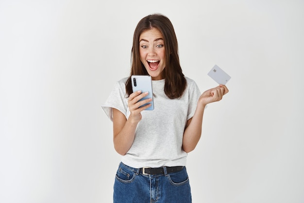 Compras en línea y comercio electrónico Mujer joven feliz usando teléfono móvil y sosteniendo una tarjeta de crédito plástica haciendo una compra de pie sobre fondo blanco